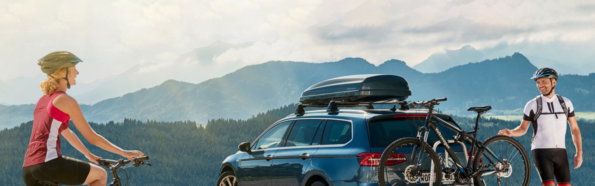 Two bikers standing beside their volkswagen with a bike mounted on the back and a roof box on the roof.