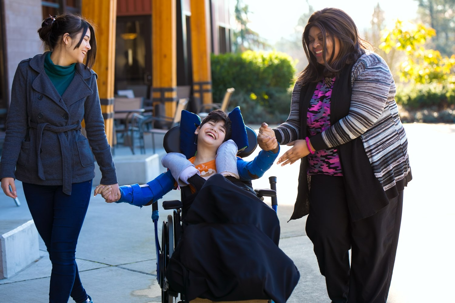 Child using a wheelchair smiling while walking outdoors with two accompanying adults.