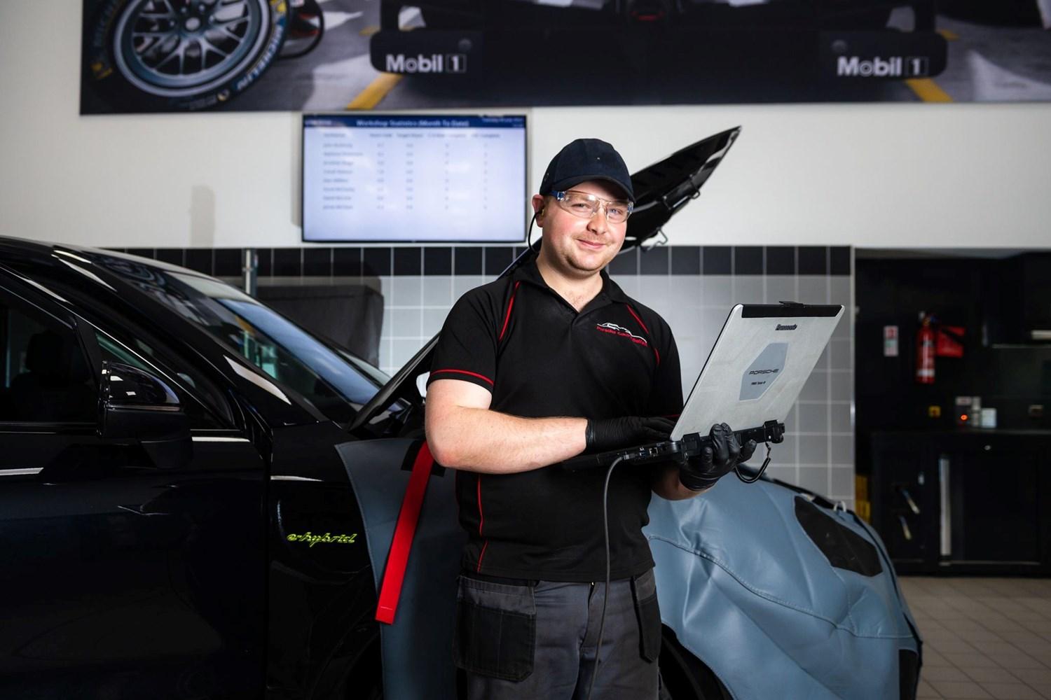 Smiling Porsche Repair Centre Repair Specialist holds laptop that is connected to the Porsche vehicle to inspect electric engine at the Porsche Approved Repair Centre at Porsche Centre Belfast