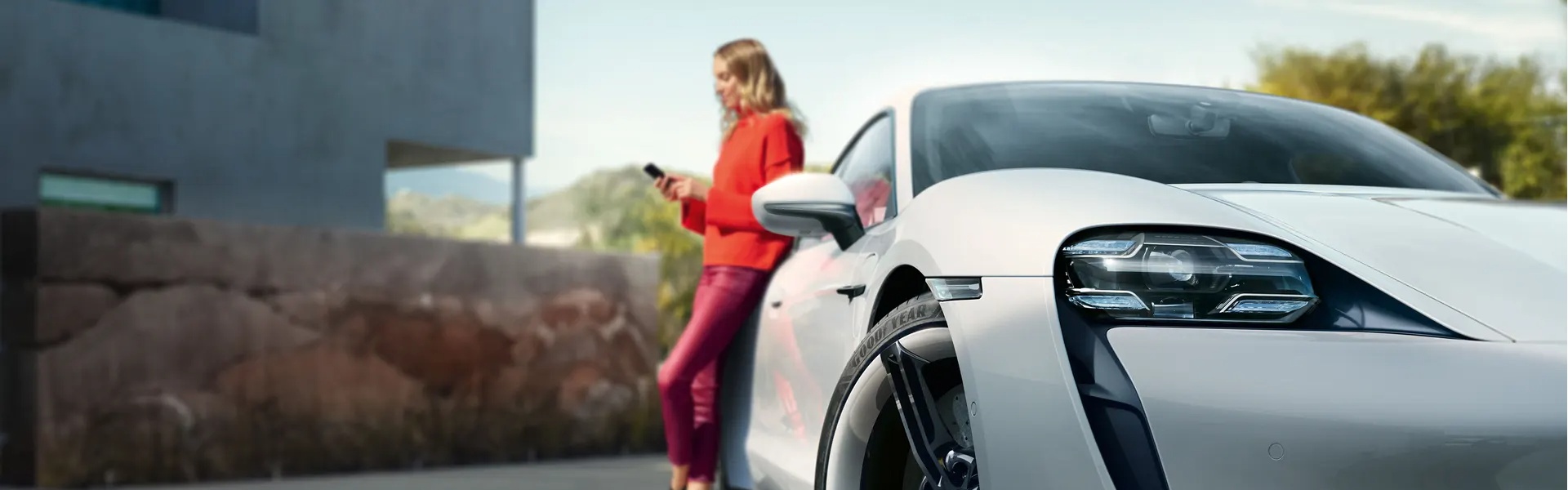 Person texting on phone leans against the rear of their white Porsche Taycan parked outside of their home