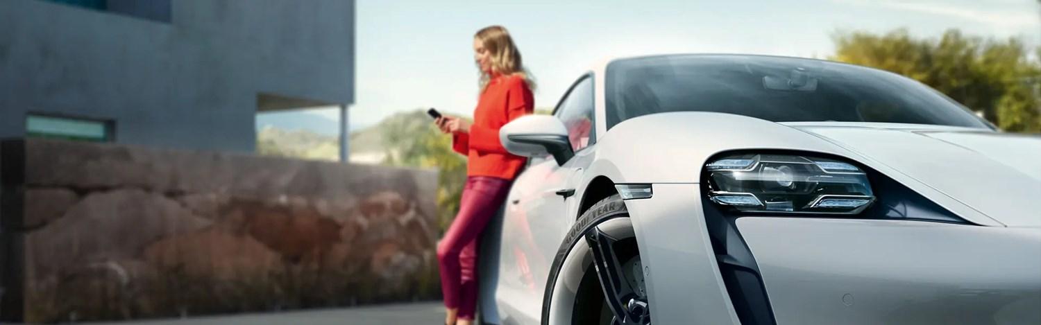 Person texting on phone leans against the rear of their white Porsche Taycan parked outside of their home