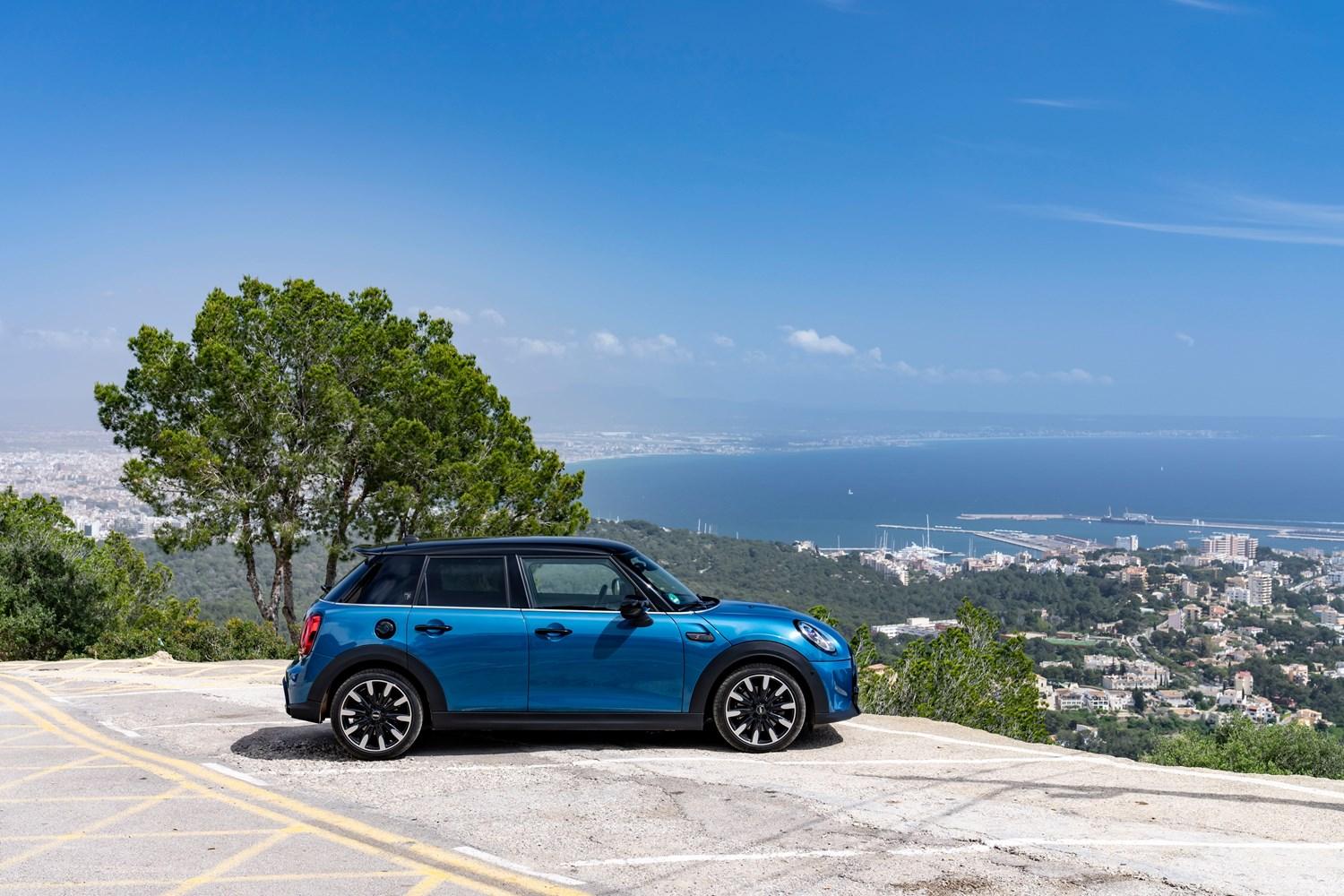 Side view of the new MINI 5-Door Hatchback in blue, parked in a car park overlooking Mediterranean city and sea