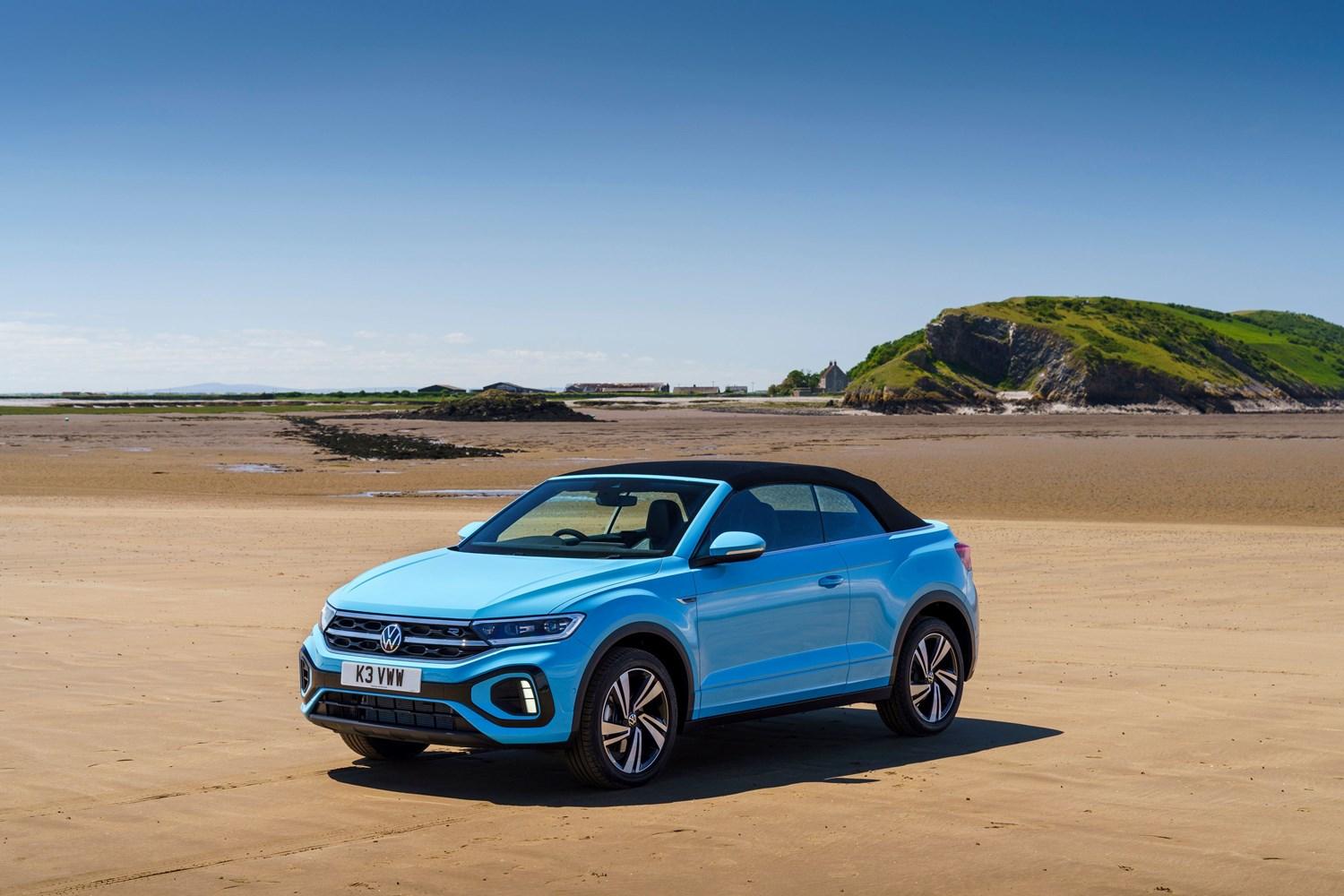 Side view of the new Volkswagen T-Roc Cabriolet in blue, parked on beach with green hills behind