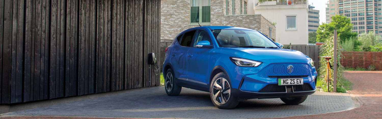 Three quarter view of a Brand new blue MG ZS EV parked in front of a brown building on a sunny day