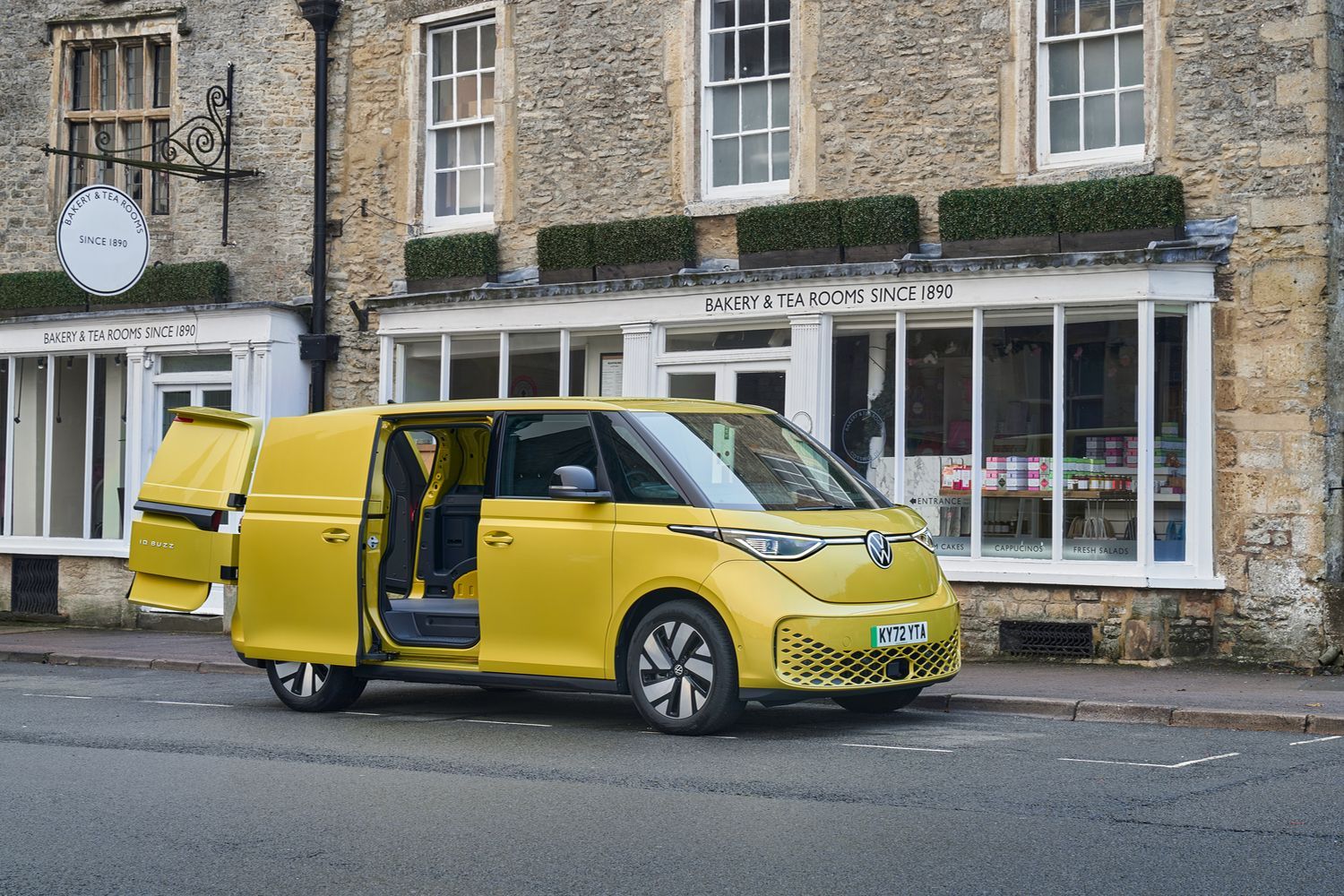 Side view of the new Volkswagen ID.Buzz Cargo in yellow, parked to the side of town street with doors open as passenger unloads packages for bakery and tearoom.