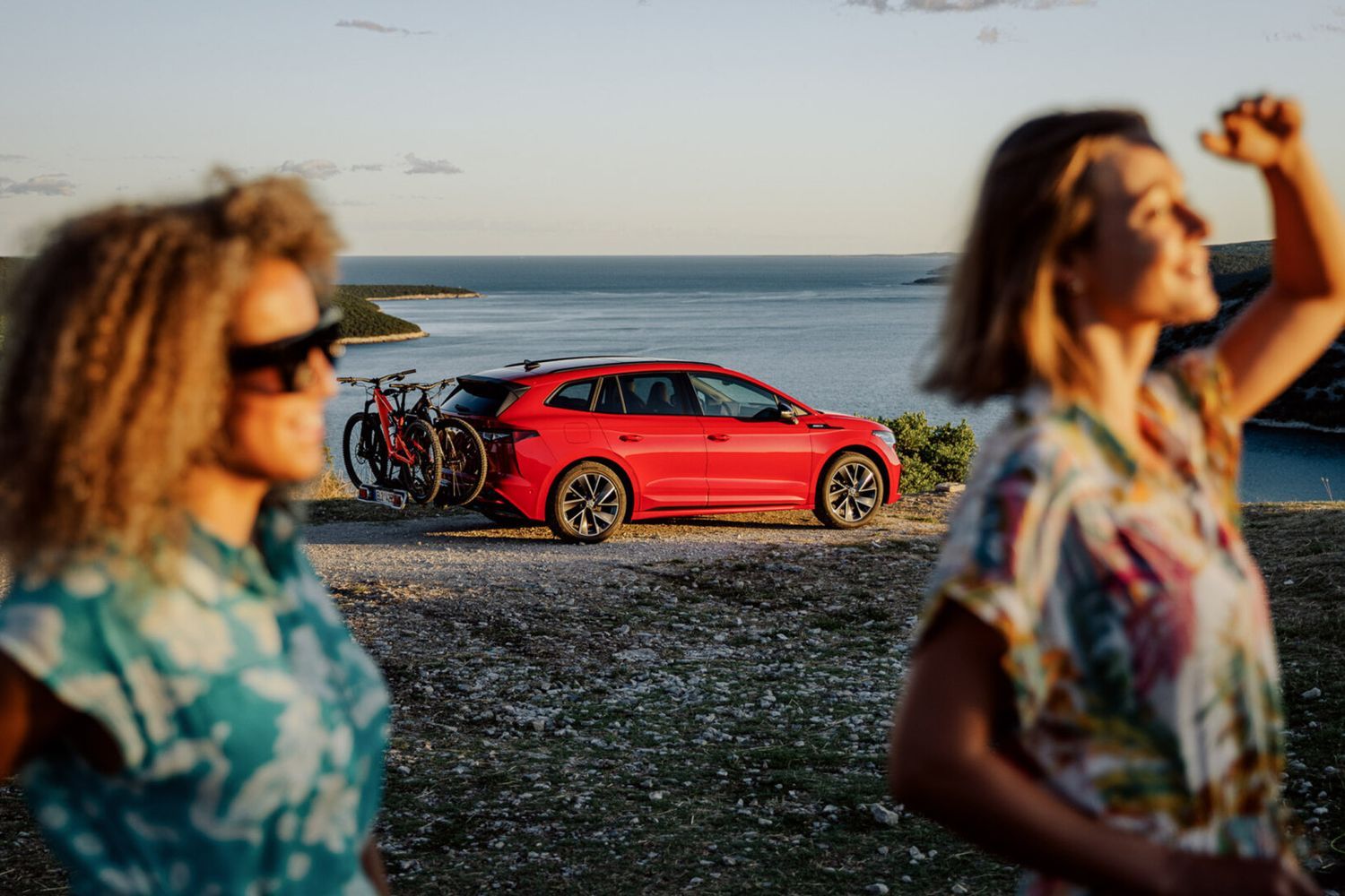 Experience Skoda. Two friends watch sunset on beach with red Skoda behind them.