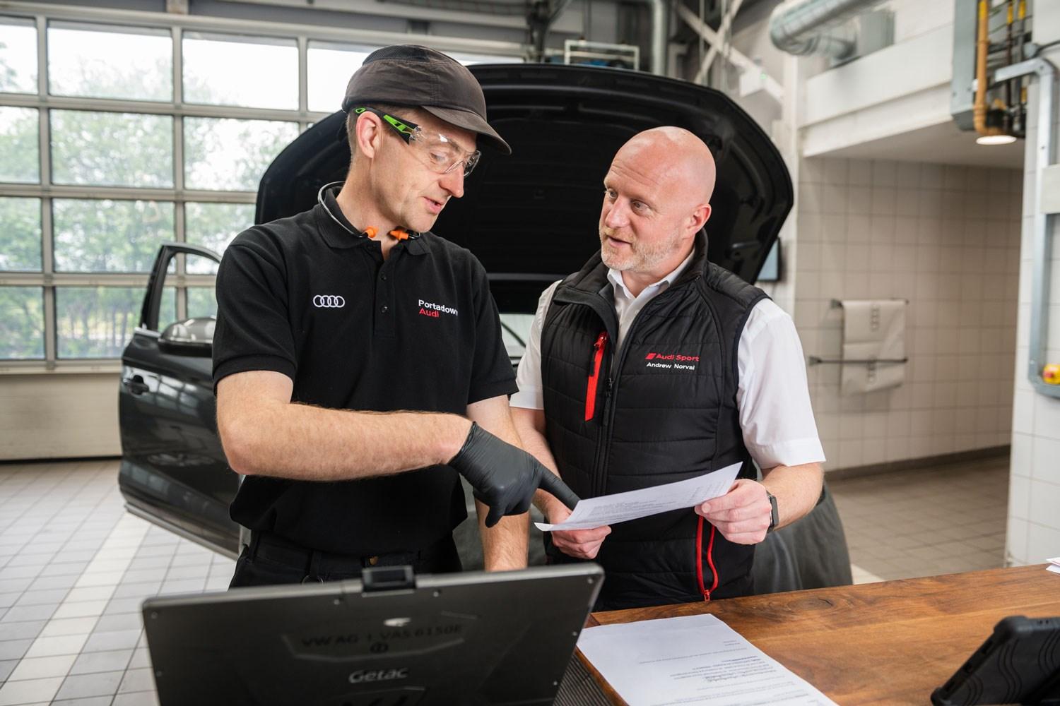 Two Audi Repair Specialists point at paper copy of warranty as they discuss repairs needed on the car behind