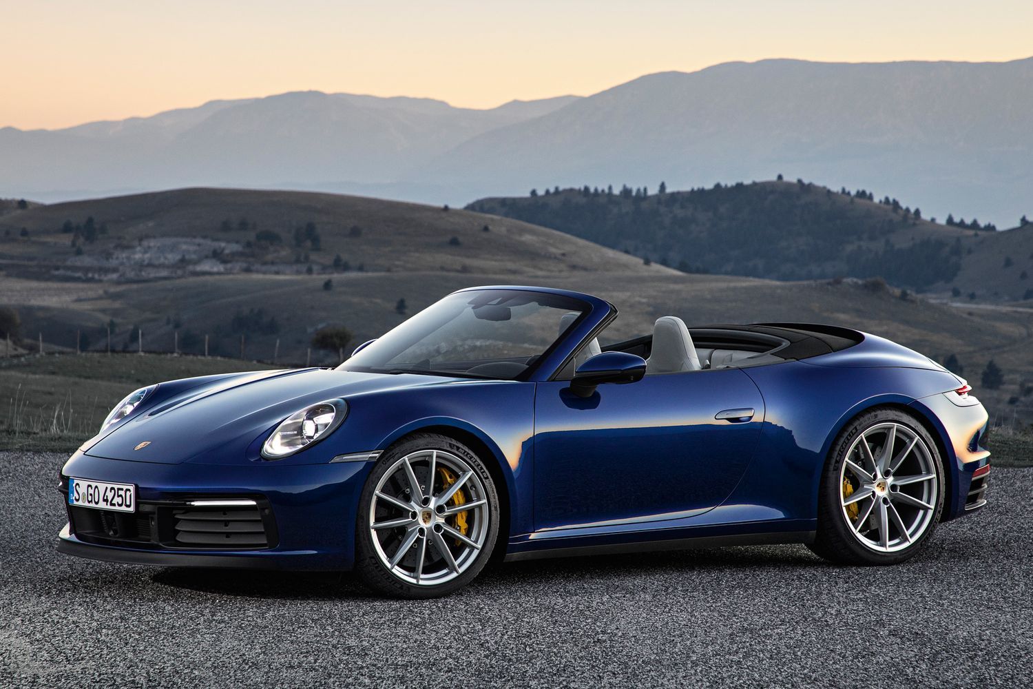 Side view of the all-new Porsche 911 Cabriolet in blue, parked in countryside with hills behind