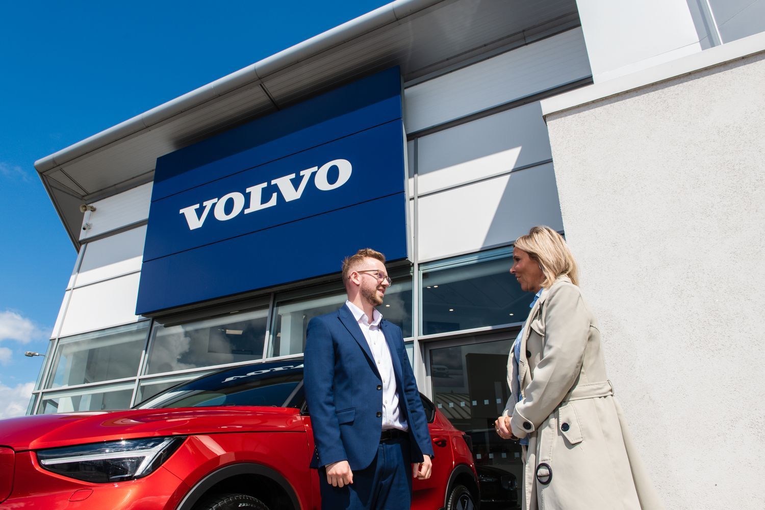 New Car Sales Specialist at Agnew Volvo Belfast chats to customer outside the showroom about the latest new red Volvo XC40 Recharge that they are standing beside