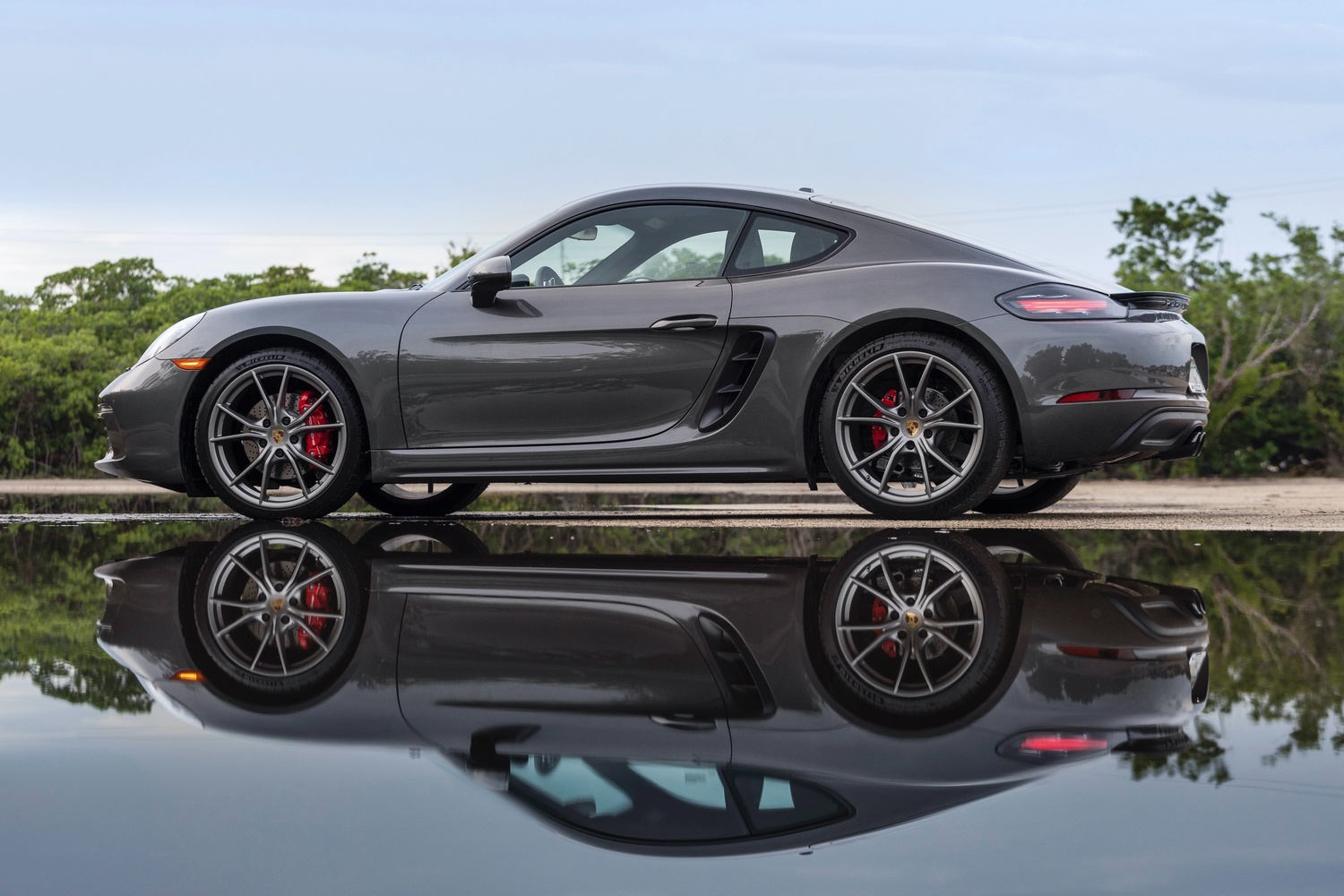 Side view of the new Porsche 718 in grey, parked on country lane with reflection in large puddle