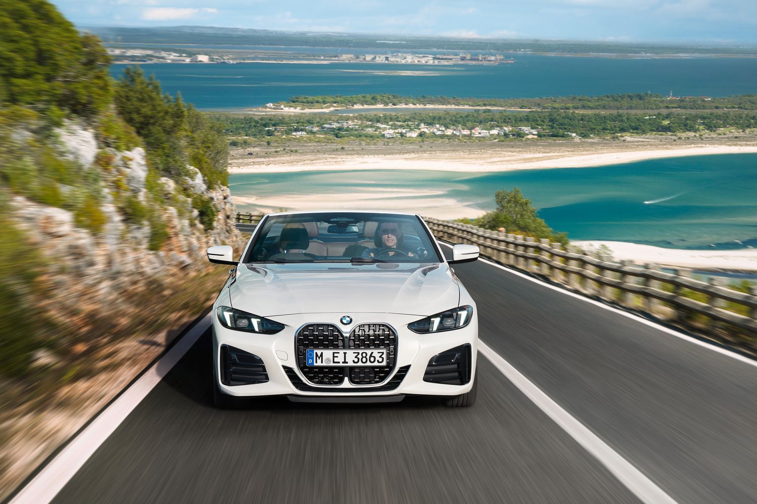 Front view of the BMW 4-Series Convertible, in white, driving up hill with beach and sea behind