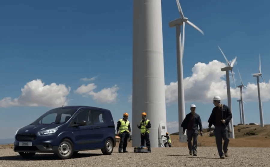Blue Ford Vehicle front side view in front of wind turbines with people stood next to it