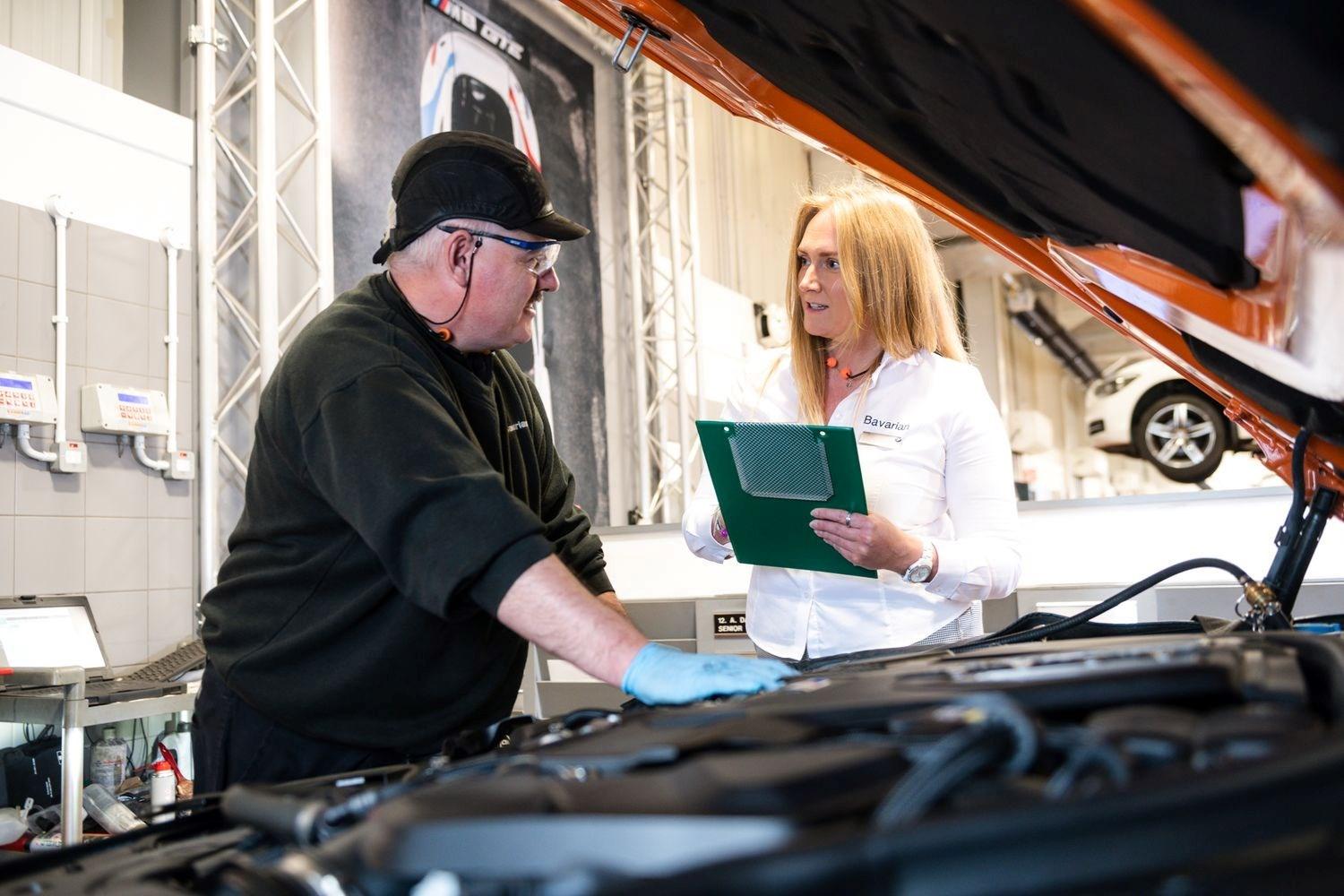 BMW Service Specialist holding clipboard and BMW Technician who is inspecting under the hood of a BMW vehicle, discuss the repairs needing made to a BMW vehicle at the Bavarian BMW Belfast repair centre.