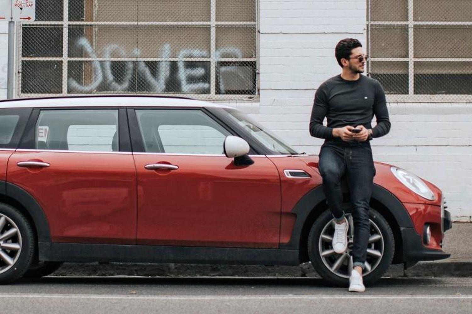 Person stands against front wheel of MINI 5-Door Hatchback while holding phone and looking away from the vehicle