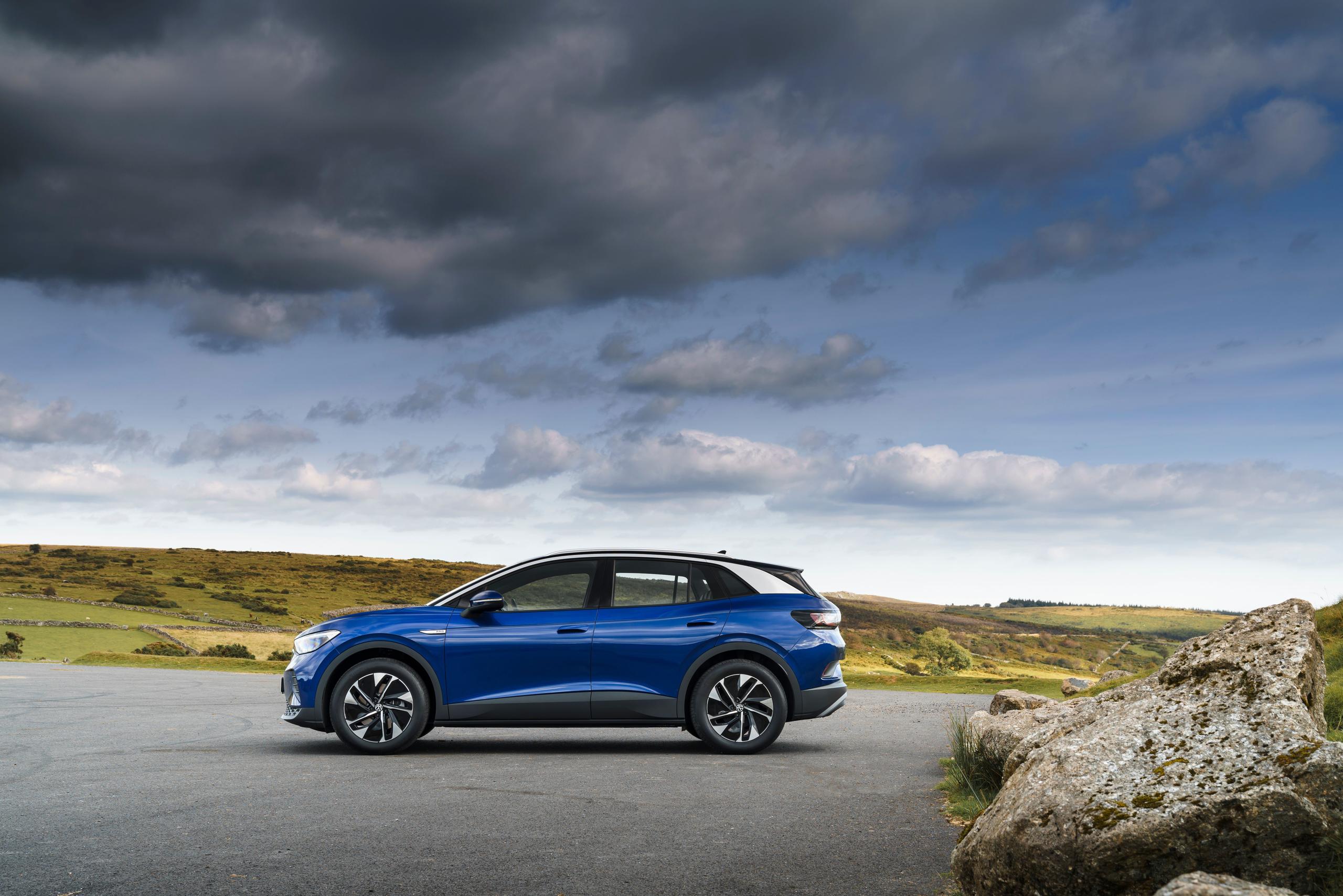 Side view of the new Volkswagen ID.4 in blue, parked in a country car pack with green hills and rocks behind
