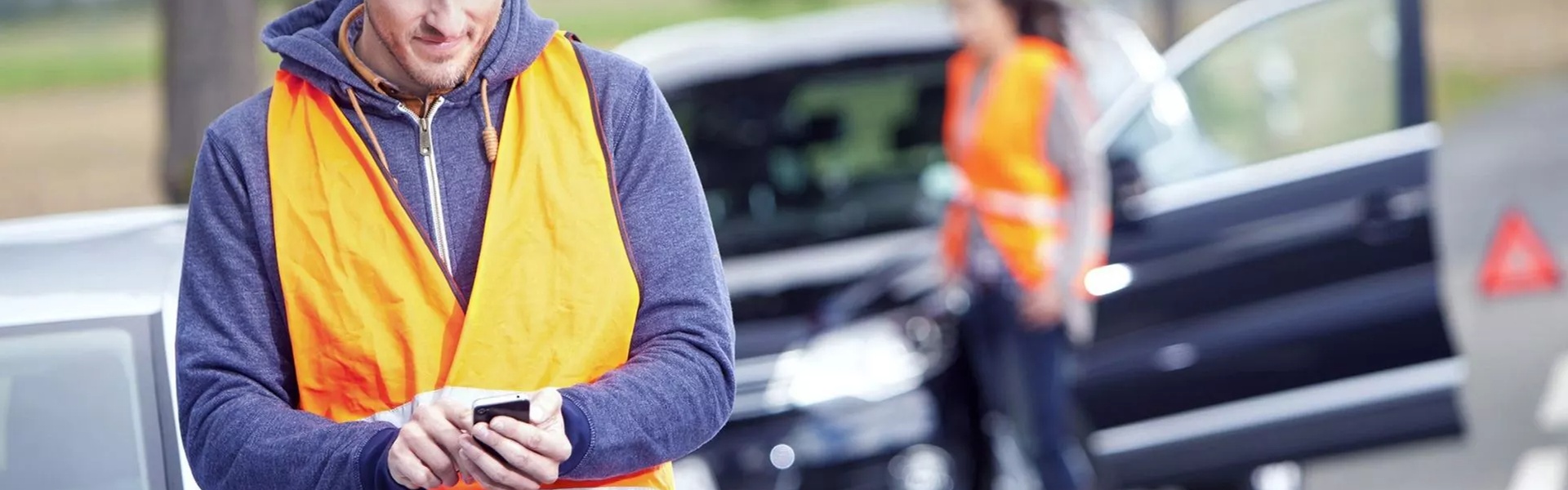 Volkswagen customer awaits roadside assistance for his Volkswagen Touran. They are currently sending a message on their mobile phone, whilst wearing a hi-visibility jacket. A warning triangle and passenger wearing a high-visibility jacket are in the background.