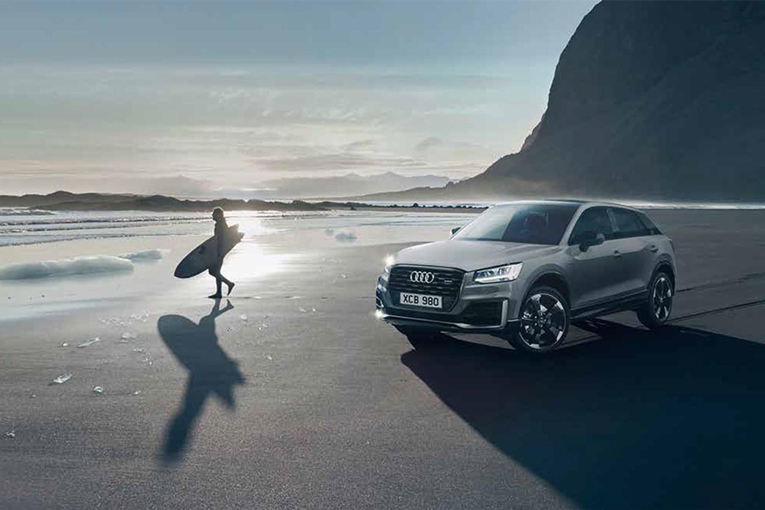 Audi Q5 is parked on a sandy beach with person walking towards sea holding a surf board