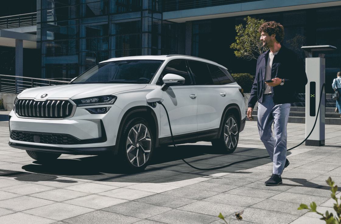 Three quarter view of a moon white metallic Skoda Kodiaq getting charged by a charging station, visible in the picture. A men walking past the car, wearing light blue trousers and and a dark blue jacket, holding a mobile device in his left hand.