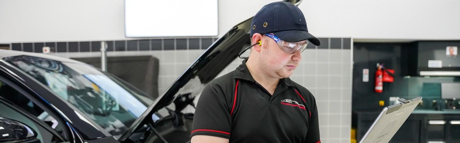 Porsche Repair Specialist reviews laptop for repairs as Porsche vehicle is behind with the hood raised.