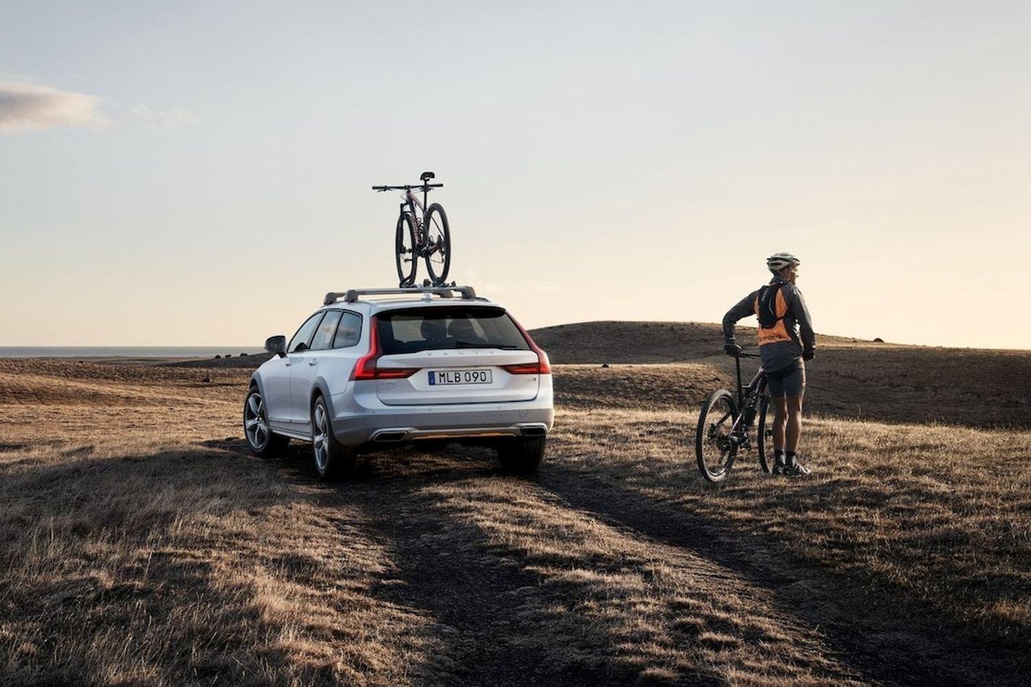 Volvo XC90 Recharge parked in field with roof cycle pack installed with bicycle loaded. To the right of the vehicle, is the passenger with bicycle.