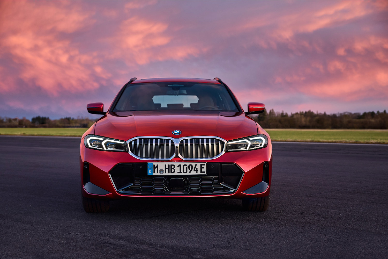Front view of the newly updated 2024 BMW 3 Series Touring in red, in front of a pink sky.