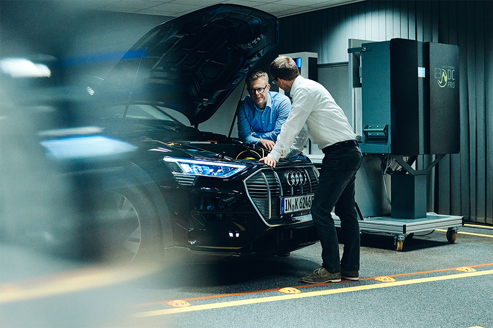 Two people stand beside an Audi Q2 with the hood up that has broken down in a car park