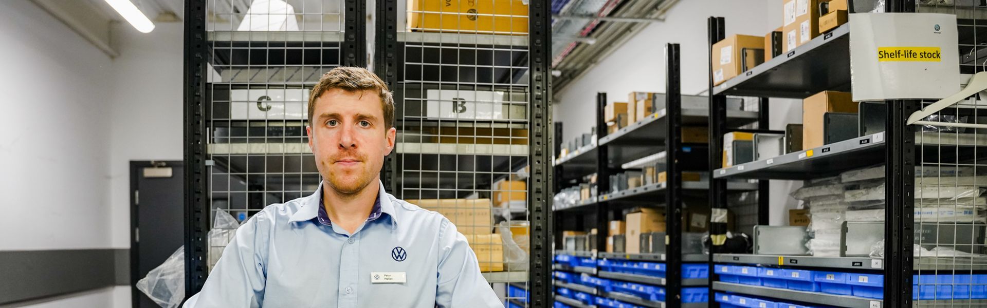 Volkswagen Parts Specialist stands behind counter with alphabetised parts stored on shelves behind.