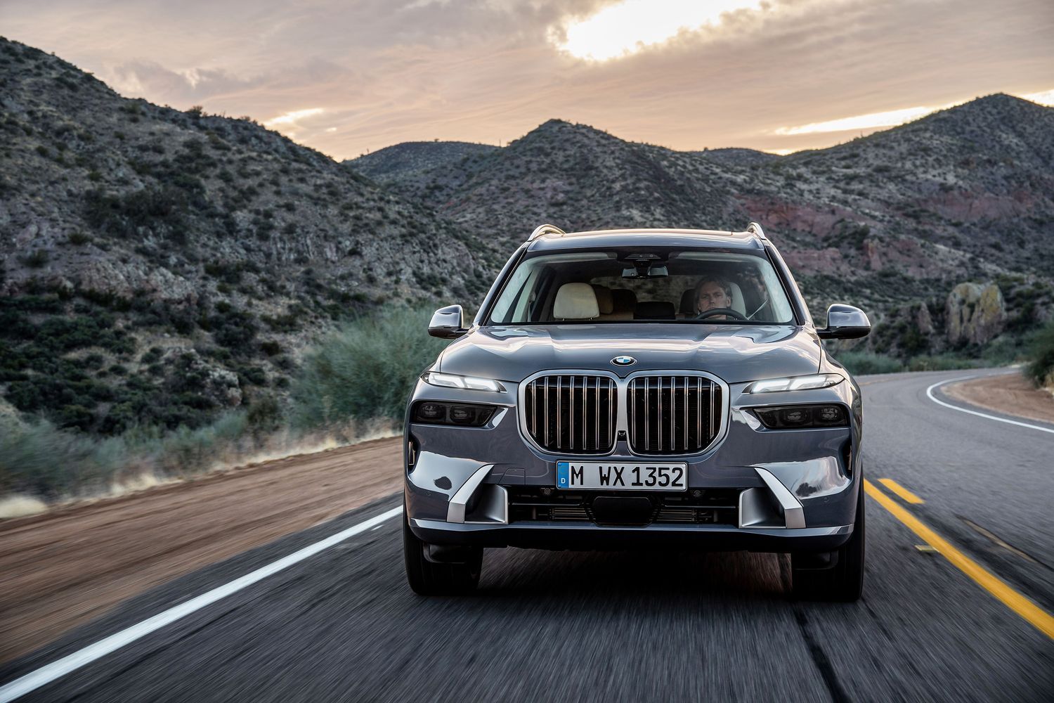 Front view of new BMW X7, in grey, driving along countryside road with mountains in background