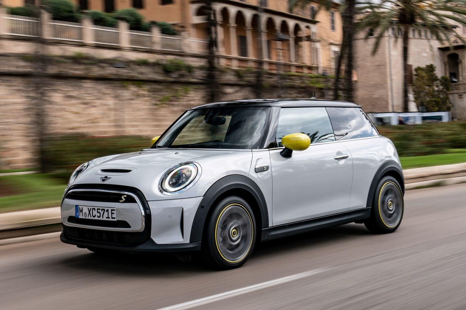 Exterior view of the new MINI Electric in white, driving along Mediterranean city with palm trees behind