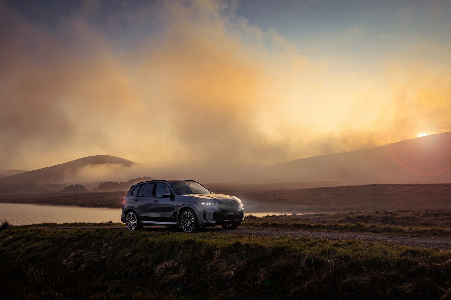 Side view of the BMW X2, parked by foggy lake and mountain view as the sunrises.