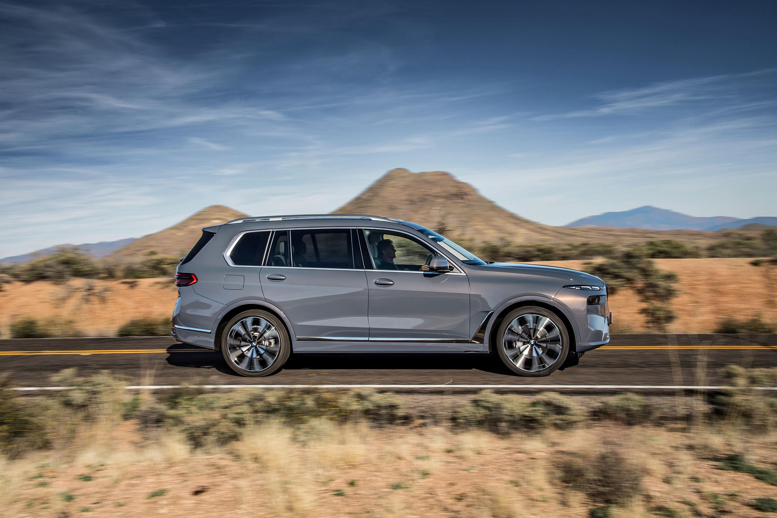 Side view of new BMW X7, in grey, driving along desert countryside road with mountains in background