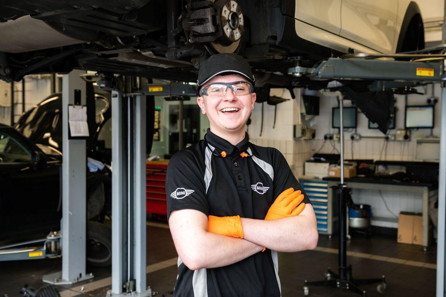 Smiling MINI Repair Specialist poses with crossed arms at the Bavarian MINI Repair Centre with MINI vehicle lifted in the background