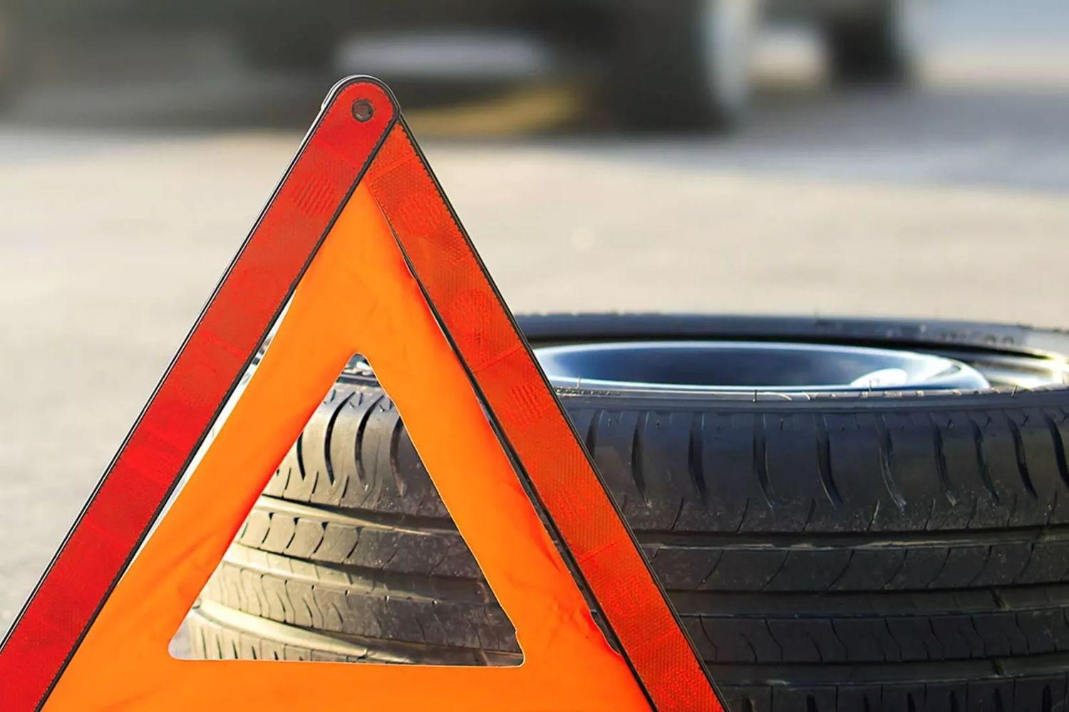 Close-up of vehicle warning triangle in front of spare tire lying flat on the road with vehicle in background.