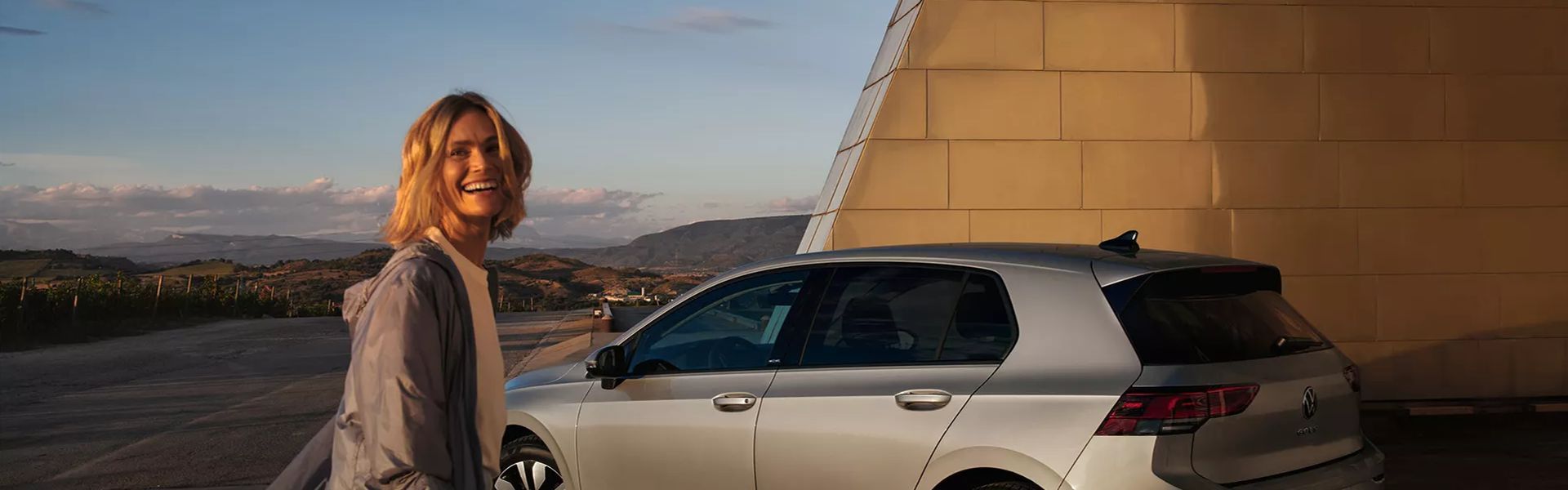 Person smiles in front of a silver Volkswagen Golf parked at the side of the road with countryside hills in the background.