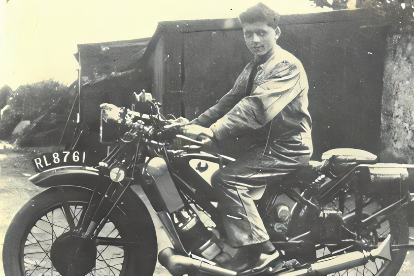 Historic black-and-white photograph of a man seated on a vintage motorcycle outdoors.