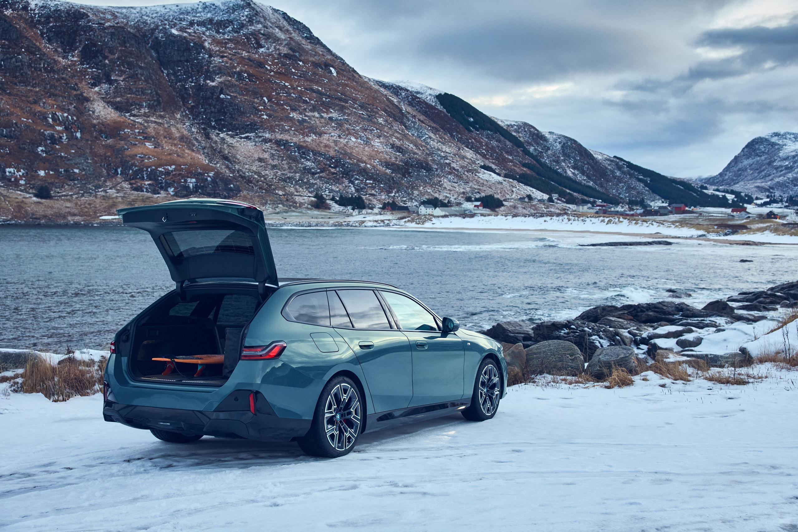 New BMW i5 Touring in green, parked in front of ice covered lake with rear boot door open and surf board visible