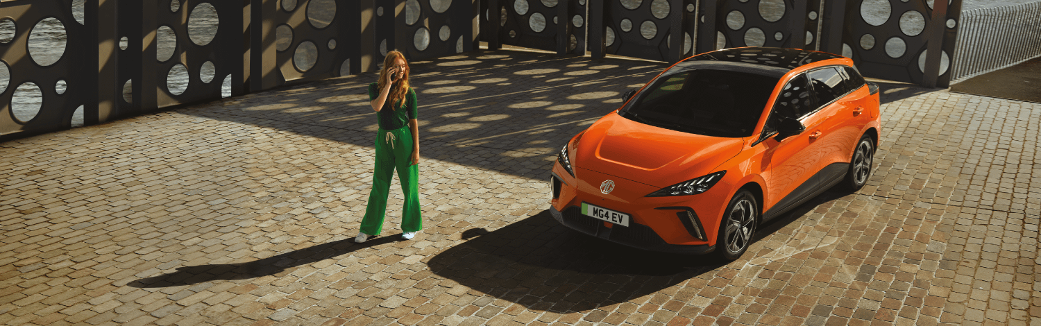 Top three quarter view of an Orange MG4 EV and a young ginger woman speaking on a smartphone outdoors in front of an artistic metal wall with round holes in it on a sunny day