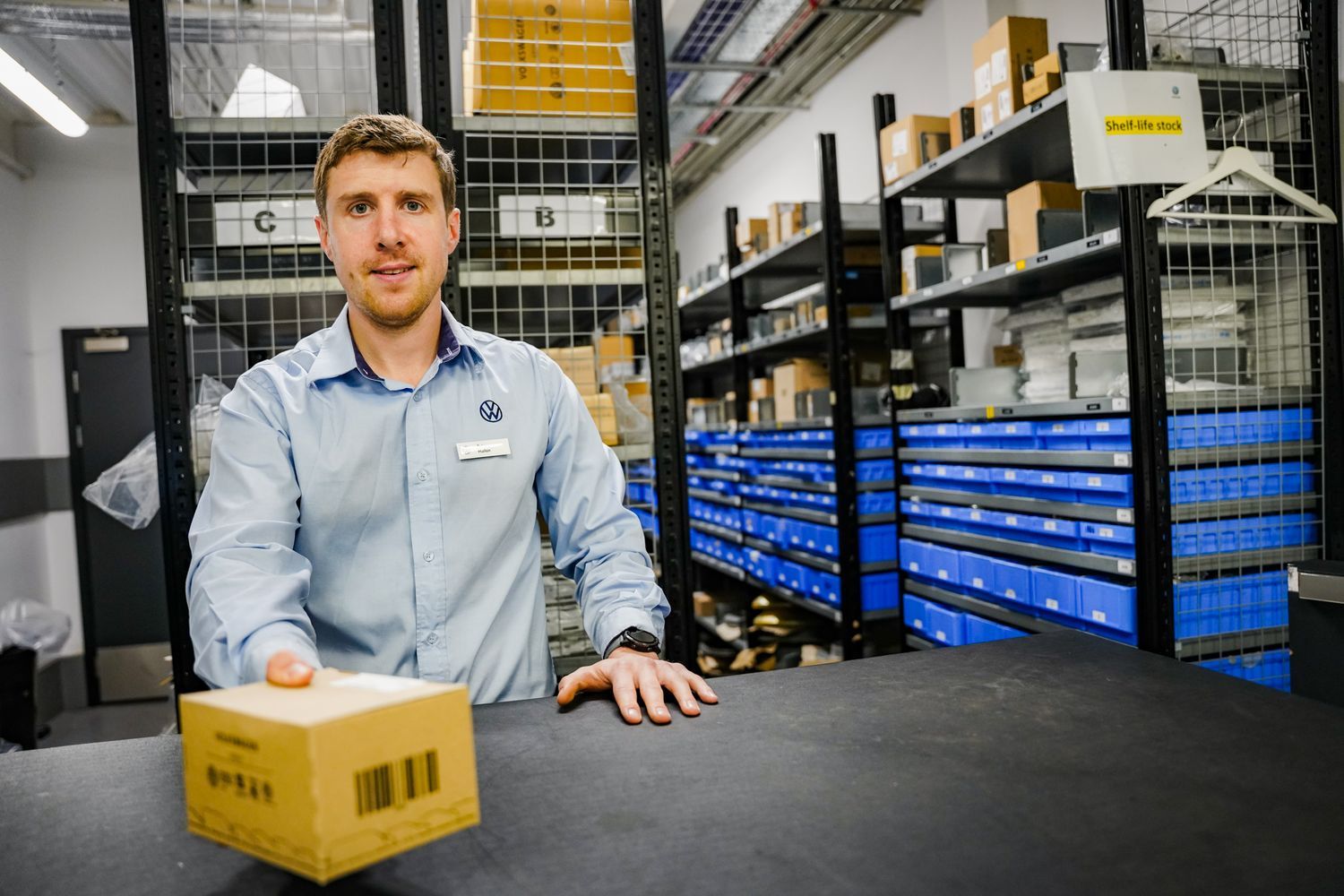 Volkswagen Parts Specialist hands over parts to customer behind counter with alphabetised parts stored on shelves behind.
