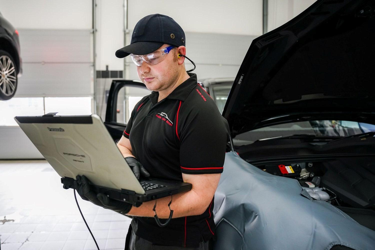 Porsche Repair Specialist reviews laptop for repairs as Porsche vehicle is behind with the hood raised.