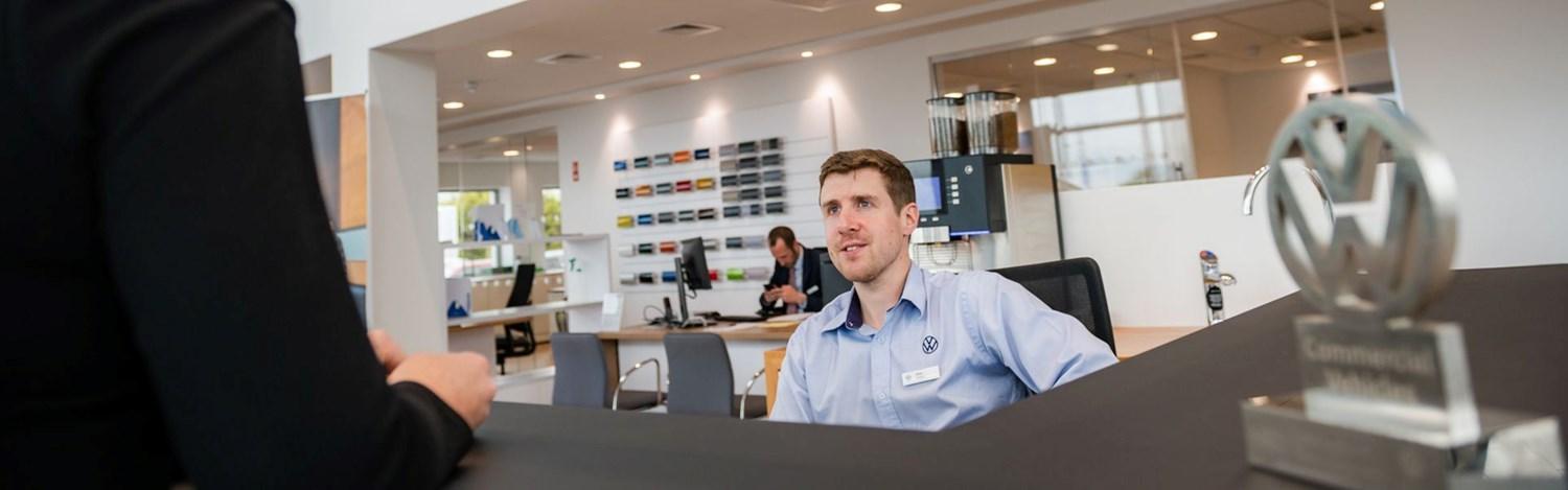 Volkswagen Sales Advisor greets customer at desk at Agnew Volkswagen Belfast, Northern Ireland. Located on the desk, is the No.1 Fleet Business Partner in the UK 2022 award that was won by the dealership.