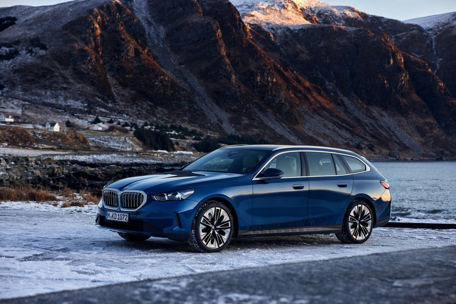 Side view of the new BMW 5-Series Touring in blue, parked by snow covered lake and mountains