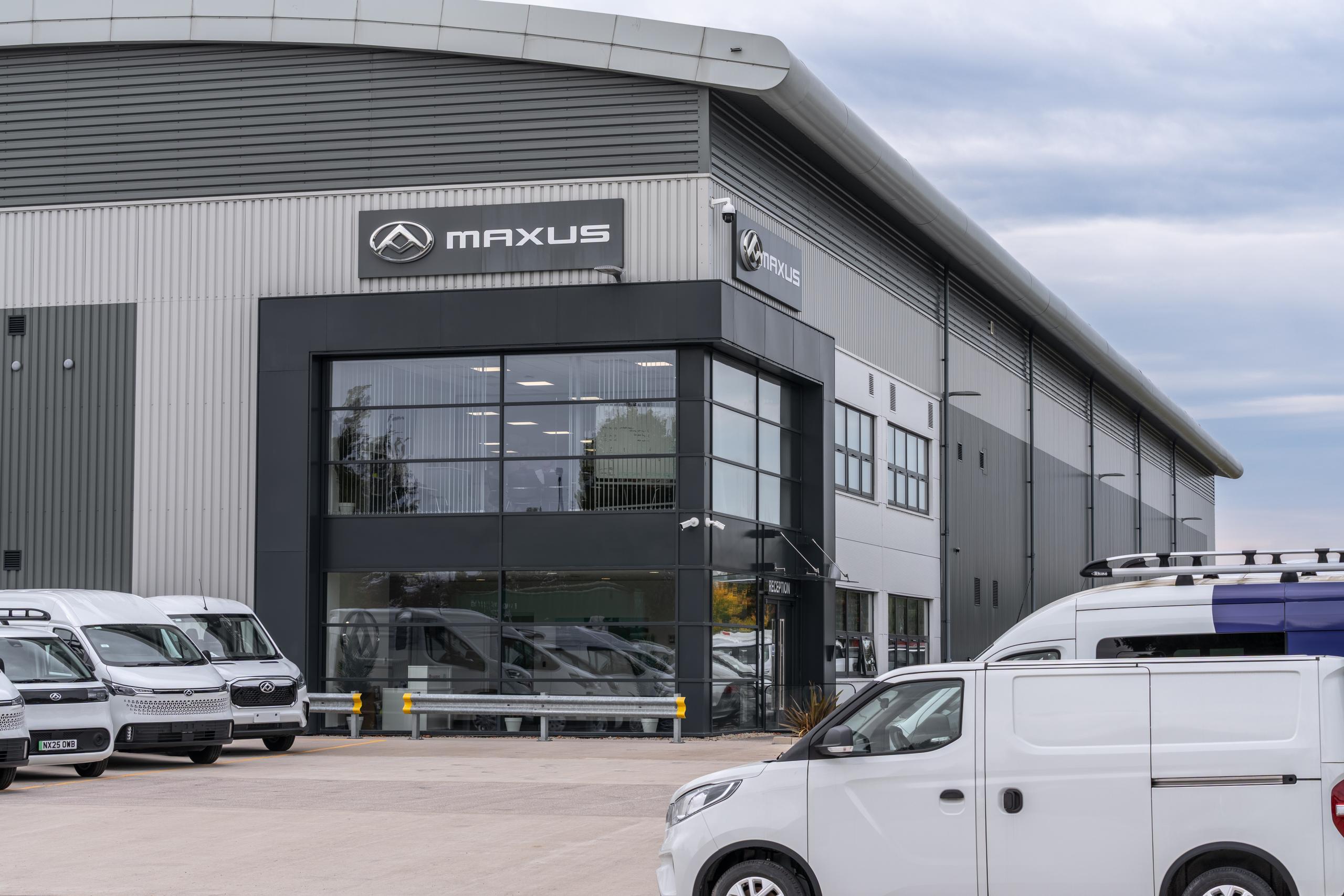 Maxus dealership building with glass entrance and several vans parked outside.